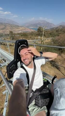 A man with short brown hair, a beard, and tattoos on his arms smiles and touches his hair as he sits in a black seat with a harness. He is wearing a white t-shirt and light-colored shorts. He has a black bag with a checkered pattern on his lap. The seat is attached to a metal track that winds through a mountainous landscape with a body of water in the distance. The video cuts to a shot of the track extending upwards into the sky, then to a shot of a person's legs and feet from a first-person perspective as they sit in a similar seat on the track. The camera then pans up to show a hand making a peace sign gesture. The scene appears to be a scenic mountain toboggan run on a clear, sunny day.