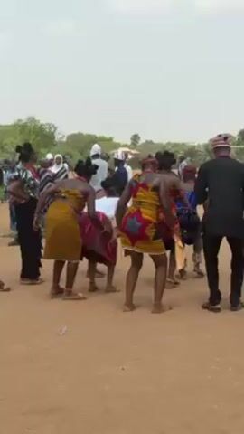 In an outdoor setting, a group of people are gathered, with several individuals engaged in a traditional dance. The dancers, predominantly women, are adorned in colorful, patterned dresses and have their hair styled in neat buns.  They also wear what looks to be coral necklaces and anklets. Their movements involve bending at the waist, with their backs to the camera. To the right of the dancers, there is a line of men playing musical instruments, including drums and a type of jug.  Some of the men are wearing traditional caps and attire.  The setting appears to be an open dirt field, and in the background, there are trees and other spectators. The day is bright, suggesting it is daytime.