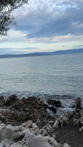 The video shows a rocky shoreline with the ocean in the foreground. Waves are gently crashing against the rocks. In the distance, there is a hazy mountain range under a cloudy sky. A few branches of a pine tree are visible in the upper right corner of the frame. The overall scene is serene and natural, with muted colors suggesting either early morning or late evening light. There are no people or man-made objects visible in the frame. The camera appears to be stationary, capturing a fixed perspective of the seascape.