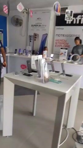 The video shows the interior of what appears to be a mobile phone store. Multiple display tables are stocked with devices. There are promotional posters for various brands of phones such as Infinix, Redmi, and Honor. Several people are present in the store, including customers browsing and staff assisting them. The camera initially focuses on an employee wearing an "Honor" t-shirt before panning around the space. The floor is tiled, and an electric fan is running to cool the space.