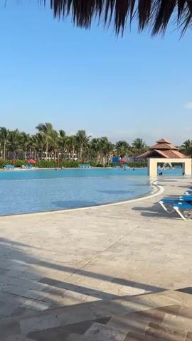 The video shows a resort pool area. The pool is large and blue with lounge chairs and umbrellas around it. There are palm trees and buildings in the background. A large sign that says "I LOVE PUNTA SAL" is visible in the distance. The sky is clear and blue. The video is filmed from under a thatched roof structure. There are no people clearly visible in the video, though there may be some figures in the distance in the pool.