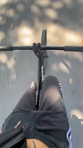 The video shows a first-person view of someone riding a black bicycle on a cracked asphalt road. The rider is wearing black pants with white stripes. There is a small black bag attached to the rider's waist.  The sun is shining, casting shadows from trees on the road. There are houses and green grass on either side of the road. A car is driving in the distance.