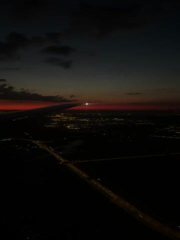 The video shows a view from an airplane at dusk. The sun is setting on the horizon, casting a red glow across the sky. Below, a city is visible, its lights twinkling in the darkness. A highway can be seen stretching across the landscape, illuminated by streetlights. The wing of the airplane is visible in the foreground, casting a shadow over the scene. The clouds are dark and scattered, adding to the dramatic effect of the twilight sky. The overall mood is one of tranquility and awe, as the viewer witnesses the transition from day to night from a unique vantage point.