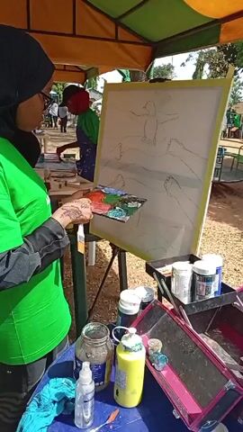 Under a tent-like structure, a woman wearing a black hijab, glasses, and a green shirt stands in front of an easel. She holds a paint palette in her left hand and a paintbrush in her right. The canvas on the easel displays a sketch of a bird surrounded by what appear to be human arms. To her right, a pink makeup box is open, revealing bottles and containers of paint. A table beside her holds more paint supplies, including a jar, a yellow bottle, and a blue cloth. In the background, another person is painting, and there are glimpses of other people and park-like scenery.