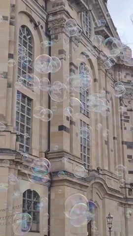 The video shows a building with large arched windows, possibly a church or an old government building. The facade is made of stone blocks. Numerous soap bubbles are floating in front of the building, obscuring parts of the windows and the architectural details. The bubbles are of various sizes and reflect light, showing iridescent colors. The sky appears overcast. There are no people visible in the frame. The camera appears to be focused on the building and the bubbles.