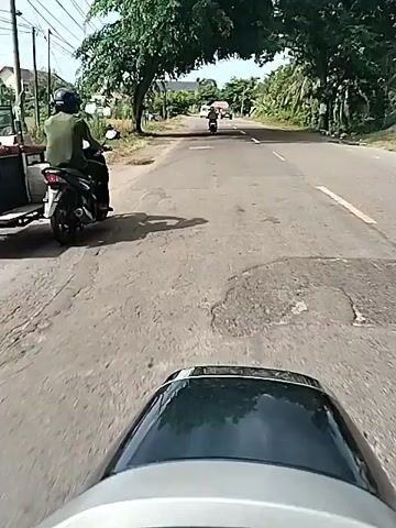 A first-person view from a moving scooter shows a person riding along a paved road in Aluekeujruen, Indonesia. The road appears to have some uneven patches. On the left side of the road, there is an open field with power lines and utility poles, and some buildings are visible in the distance. On the right side, there are dense green trees and foliage, including palm trees. The sky is partly cloudy and bright, indicating daytime. In the distance, other vehicles, including a motorbike and a black SUV, are seen approaching or traveling on the road. The scooter's dashboard/windshield is visible at the bottom of the frame. Wind noise is audible, suggesting movement. No visible text is present.