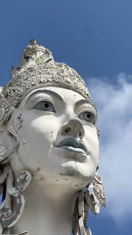 The video shows a close-up of a large, white statue against a blue sky with scattered clouds. The statue appears to be of a deity, possibly a Hindu goddess, with intricate headwear. The statue's face has blue lips and eyes. The statue has some weathering. The camera angle looks up at the statue. The background consists of the clear blue sky, with intermittent clouds.