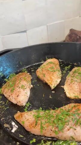 A cast iron skillet is on a stovetop, filled with four pieces of seasoned chicken breast. The chicken is garnished with chopped green herbs, likely parsley. The chicken is in the process of being cooked, with some browning visible on the edges. In the background, there are multiple steaks on a separate cooking surface, possibly another pan or grill, also appearing to be cooked. The kitchen backsplash is made of white, textured tiles. The lighting suggests an indoor setting, likely a kitchen.