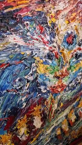 The video features a close-up, static shot of a highly textured, abstract painting of a cat's face. The painting utilizes a heavy impasto technique, with thick, raised applications of vibrant oil paint in various colors including blue, yellow, red, green, and white. The cat's eyes, nose, and mouth are discernable through contrasting colors and textures. The background and surrounding areas of the canvas are filled with energetic, abstract strokes of similar bright colors. The camera slowly zooms out over the duration of the video, revealing more of the artwork's full composition.