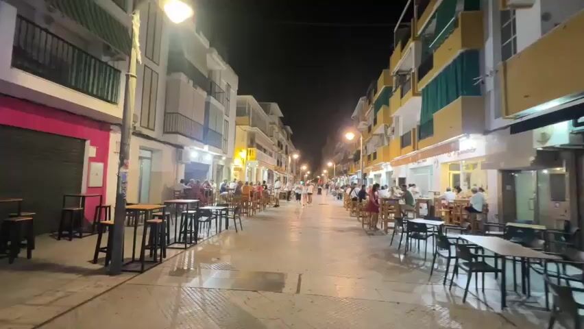 The video shows a pedestrian street at night. Buildings with multiple floors line both sides of the street. The buildings have balconies and windows. There are many tables and chairs set up outside restaurants and bars on both sides of the street. People are sitting at the tables, eating and drinking. Some people are walking down the street. The street is illuminated by streetlights and lights from the buildings. The ground appears to be wet, possibly from recent rain or cleaning. There are a few signs visible on the buildings, but they are not clearly legible. The overall atmosphere appears to be lively and social.