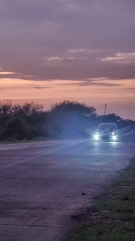 A car with its headlights on drives down a paved road during a sunset. The sky is a gradient of purple and orange. The car is dark-colored and appears to be a sedan. Trees and foliage are visible along the side of the road. The road surface is wet, reflecting the headlights.