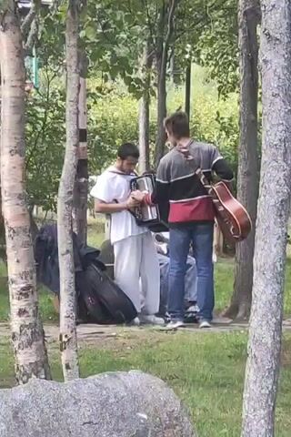 The video shows two young men in an outdoor, wooded setting. The man on the left, wearing a white tunic and white pants, is playing an accordion. He is standing and his posture suggests he is actively engaged with the instrument. To his right, the second man, wearing a dark and red patterned shirt and blue jeans, has an acoustic guitar strapped across his back. He is facing the accordion player and appears to be listening or observing. Both men are surrounded by trees and foliage, with a large rock in the foreground. There are also cases, likely for musical instruments, visible on the ground near the accordion player. The overall impression is of a casual musical moment taking place in a natural environment.