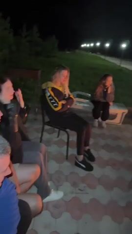 The video shows a group of people gathered around a bonfire at night. The ground is paved with interlocking bricks. In the foreground, a person wearing a black hoodie and a blue baseball cap is looking down. Behind them, a woman with dark hair is visible. Further back, several people are seated on chairs around a bonfire. One person in a yellow and black jacket is seated on a chair on the left side of the screen. Another person in a gray outfit appears to be dancing or posing in the center. In the background, there are some buildings and lights. The atmosphere appears to be relaxed and social.