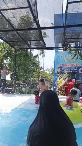 A group of people are enjoying a bubble pool. In the foreground, a man is holding a young child in his arms, both submerged in the bubbly water. To the right, a person is floating on a yellow inflatable ring. In the middle ground, a child in a green shirt stands in the bubbles, looking towards the left. Further back, another child in a pink inflatable ring sits in the water, with a person in a white shirt standing behind them, seemingly playing. On the left side of the frame, a person in a white shirt and dark pants stands on a raised platform, holding a stick and appearing to be throwing something. Behind them, trees and foliage are visible. A blue structure with text that appears to be a sign is situated in the background. The environment is outdoors, under a black mesh canopy.