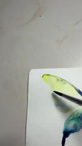 A close-up video shows a paintbrush with a silver ferrule and black handle being used to apply watercolor paint to a white piece of paper. The paint is a vibrant yellow and green, with hints of blue. The paper is on a light gray surface, possibly a table, which has some water stains on it. The painting appears to be abstract, with flowing lines and blended colors. The brush strokes are delicate and precise, creating a soft, ethereal effect. The video is filmed from a high angle, looking down at the artwork.
