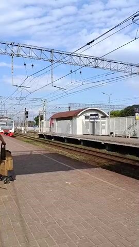 The video opens with a view of a train arriving at a station. A person is standing on the platform with a shopping cart. The train, which appears to be an electric multiple unit, has a white and grey exterior with orange stripes. The station platform is paved with bricks and has overhead power lines for the train. A small building with a brown roof and white walls is visible on the platform. As the train pulls into the station, the camera pans to show the side of the train, revealing its windows and doors. Several people can be seen inside the train through the windows. The sky is blue with some clouds.