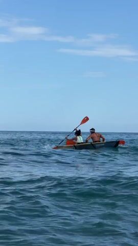 Two people are in a double kayak on the ocean under a partly cloudy sky. The person in the front is paddling with a red paddle. The person in the back is facing forward. The kayak is in motion and appears to be moving through gentle waves.