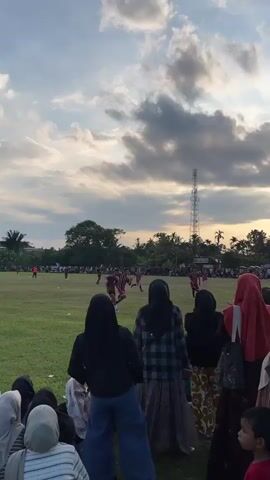 A group of people, mostly women wearing hijabs, stand on a grassy field watching a soccer game. The sun is setting, casting a warm glow across the sky, which is filled with dramatic clouds. In the distance, players can be seen on the soccer field, and a tall communication tower stands on the horizon. The atmosphere appears to be festive and communal.