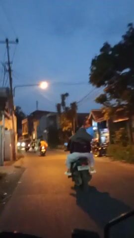The video shows a first-person perspective from someone riding a motorcycle on a street in what appears to be a residential or mixed-use area. The sky is a deep blue, indicating dusk or early night. Streetlights and vehicle headlights illuminate the road. The rider is following another person on a motorcycle, who is wearing a head covering and light-colored clothing. Other vehicles, mostly motorcycles, are visible ahead and parked on the sides of the street. Buildings line both sides of the road, and some trees are scattered along the route. The street appears to be paved and well-lit by streetlights. The overall scene suggests a typical evening commute or ride through a town in Banjaran, Indonesia.