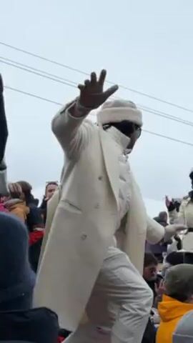 A man wearing a long, white coat and a matching fur hat dances in a crowd. He faces away from the camera, his right arm raised. Many people surround him, some holding drinks. The sky is overcast.