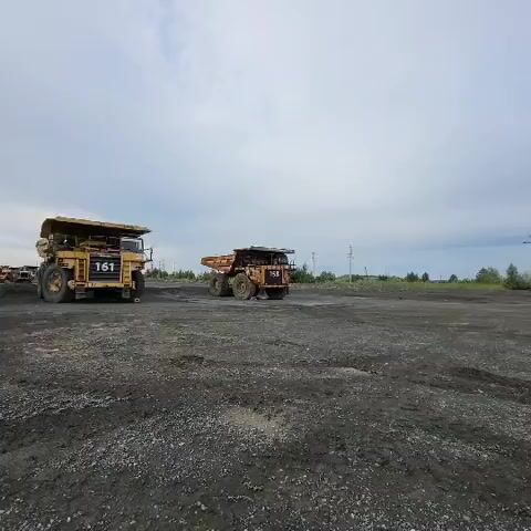 This media file captures a scene at a mine in Voloshyne, Ukraine. The primary subjects are two massive yellow Caterpillar mining trucks. The truck in the foreground, numbered 158, is the main focus, with its rear and side clearly visible. It is a dump truck designed for hauling heavy loads, evident from its large tires and robust construction. To the left, partially obscured by the camera's perspective and possibly further away, is another similar mining truck, numbered 161.

The setting is an open-pit mine, characterized by a vast expanse of dark, dusty ground, likely coal or other excavated material. The terrain appears uneven and industrial. In the background, a line of trees and shrubs suggests the edge of the mining area, with utility poles and electrical lines indicating infrastructure. The sky is overcast with muted clouds, suggesting a daytime scene with potentially stable weather, not intensely sunny or stormy.

No people are visible in the media file. The activity appears to be the static presence of these large mining vehicles, possibly parked or waiting for their next task. The overall mood is one of industrial scale and quiet operation, devoid of immediate action. The visible text includes the truck identification numbers, "158" and "161," and the brand name "CATERPILLAR" emblazoned on the front of the truck. The scene conveys a sense of the heavy machinery that operates in such environments.