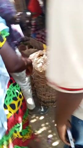 The video is shot from a first-person perspective, moving through a busy outdoor market. Initially, the camera faces towards stacked textiles and then a man wearing a plaid shirt as he moves through the stalls. The camera passes by woven rope-like materials and then the back of a person with dark braids and a white shirt. The viewpoint continues to move through the crowded area with a chaotic overhead view of the makeshift roof of the market.