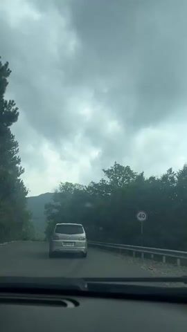 The video shows a view from the dashboard of a car driving on a paved road through a forested area. A silver hatchback car is in front of the camera car. The road curves to the left and is bordered by a metal guardrail on the right. Dense green trees line both sides of the road, with mountains visible in the distance. The sky is overcast with thick clouds. A speed limit sign indicating 40 is visible on the right side of the road. The video is filmed from inside a vehicle, suggesting the camera is also in a car.
