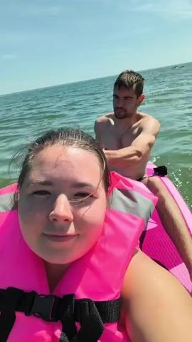 A man and a woman are on a paddleboard in the water. The woman is in the foreground, wearing a pink life vest, and holding the paddleboard. The man is in the background, shirtless, wearing black swim trunks, and paddling the board. The water is calm, and the sky is clear.