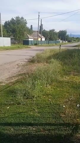 This is a video of a rural scene in Abai, Kazakhstan. The camera pans across a street with houses and fences on either side. Tall grass and trees line the roadside, and a few power poles are visible in the distance. The sky is blue with some clouds, suggesting it is daytime and the weather is fair. The overall impression is one of a quiet, rural village, perhaps in the summertime. There are no people or vehicles visible in the video, and no text is present.