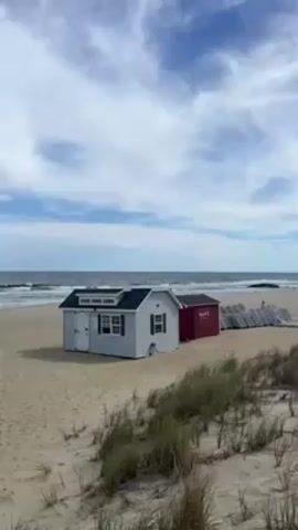 The video shows a beach scene with a small, light-colored shed and a red shed on the sand near the ocean. There are also stacked chairs and some beach grass visible. The sky is blue with scattered clouds, and the ocean is calm with small waves.