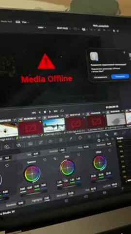 The video shows a laptop screen displaying the DaVinci Resolve video editing software. A red "Media Offline" warning is displayed in the preview window, indicating missing video files. A pop-up window in Russian appears to ask for permission to access an iPhone, likely as a source for the missing media. The timeline shows several video tracks with disconnected clips, and the color grading panel is visible at the bottom, with various color wheels and sliders. The laptop itself is sitting on a surface with a grey or beige fabric underneath. The song that is playing is in a high pitch and hard to understand.