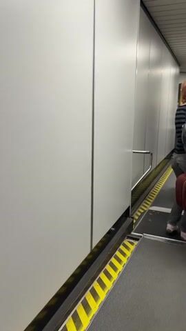 The video shows a person walking through an airport jet bridge. The jet bridge is enclosed with white walls and a yellow and black striped safety railing. The person is carrying a red suitcase and wearing jeans and a striped shirt. Other passengers can be seen further down the jet bridge. The video captures the movement of the person as they walk away from the camera and toward the aircraft.