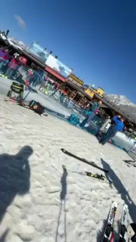 The video shows a person standing in the snow at a ski resort. The person is wearing gloves, and their skis are visible nearby. In the background, there's a building with people gathered around it. There are also other skis, snow, and a mountain range in the distance. The sky is clear and blue.