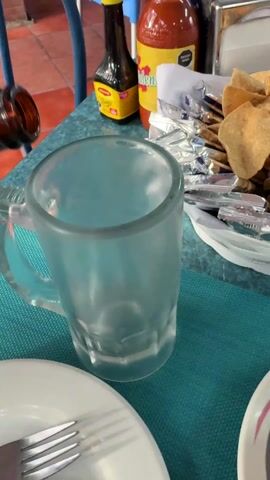 The video shows beer being poured into a frosted mug at a restaurant. A dark brown bottle is tilted and a stream of golden liquid flows into the mug, creating a foamy head. The mug sits on a teal-colored tablecloth. Beside the mug is a white plate with a fork. In the background are bottles of what appears to be Maggi seasoning and hot sauce, along with a plate of tortilla chips and packets of sauces or condiments. The floor visible in the background is made of red bricks. The restaurant appears to be outdoors.  There are sounds of people talking in the background.