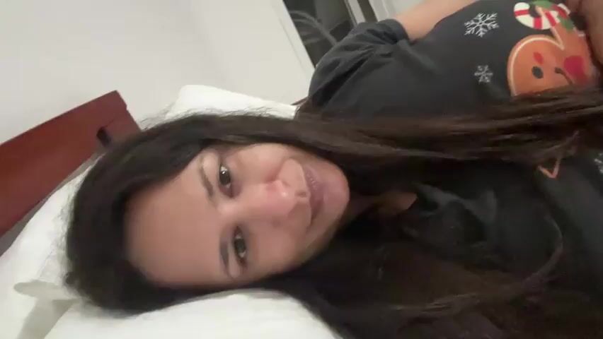 A person with long dark hair is lying in bed. They are wearing a black t-shirt with a gingerbread man and snowflakes on it. The person is smiling and making a peace sign with their fingers near their face. The bed has a white pillow and a wooden headboard.