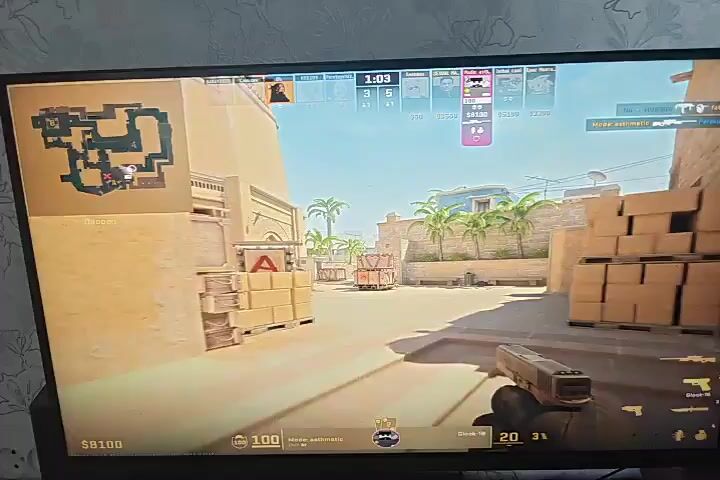 The video shows a computer monitor displaying a first-person shooter video game. The game appears to be Counter-Strike: Global Offensive, with the map "Mirage" visible on the mini-map in the top left corner. The player's perspective is from the Counter-Terrorist side, as indicated by the character model and the objective marker on the mini-map. The player is holding a knife, suggesting they are either in a stealthy situation or preparing for a close-quarters encounter. The game clock shows 1:02 remaining in the round. The environment is a desert-themed map with sandy buildings, crates, and palm trees. Other players' icons and scores are visible at the top of the screen, indicating a competitive match is in progress. The bottom of the screen shows the player's in-game money ($8100) and ammunition count. The overall scene is one of intense gameplay, with the player likely navigating the map to reach an objective or engage enemy players.