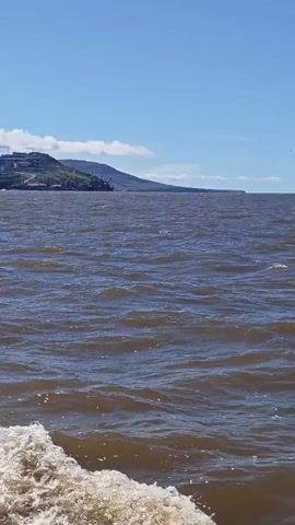 A video recording shows a view from a boat moving through dark, choppy water under a bright blue sky with scattered clouds. In the distance, a town or city is visible on a hillside overlooking the water. The town has many buildings and appears to be a port or industrial area with cranes. The camera is positioned low on the boat, capturing the white foam and wake created by the vessel's movement through the water. The railing of the boat is visible in the foreground on the left side of the frame. The video is short, lasting only a few seconds.