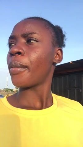 A young, dark-skinned woman wearing a yellow t-shirt is outside with clear blue skies in the background. She is speaking and looks around slightly. In the middle of the video, she looks down at the camera and puts her hand up to her forehead. There is a telephone pole with wires running up it. There is a structure made of wood and corrugated metal, and several cars are visible in the background.