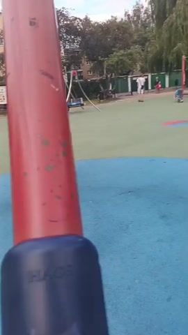 A child on a unicycle circles a metal pole in a playground. The child wears a helmet and a patterned jacket. The playground has a blue and green rubber surface. In the background, other children are playing on swings and playground equipment. Trees and buildings are visible in the distance. The sky is cloudy.