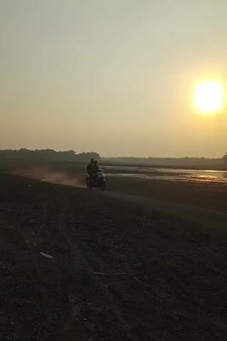 The video shows two motorcycles with riders in a dusty, open area during sunset. The sun is visible in the upper right corner, casting a warm glow. The ground is dark and appears to be dirt or gravel, with visible tire tracks. In the distance, there is a body of water and a treeline. The motorcycles are kicking up dust as they move. One motorcycle in the foreground is carrying two people.