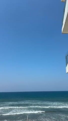 The video presents a clear daytime view of a calm ocean under a bright blue sky. Gentle waves roll towards the shore, which is partially visible in the lower frame. A faint haze hangs near the horizon. In the upper right corner of the frame, a portion of a building structure is visible, with glass railings. As the video continues, a small, distant island comes into view, along with some land to the far left. Graffiti on the wall is captured in the lower left corner of the frame. The focus remains on the ocean and sky, highlighting the peaceful coastal scene.