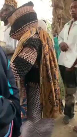 The video shows a crowd of people gathered in an outdoor area that appears to be a village or community setting.  The central figure in the video is an individual wearing a distinctive, possibly ceremonial outfit featuring woven elements and patterned fabrics. This person is the focus of the crowd's attention. The crowd is diverse, with people of varying ages and wearing a mix of casual and possibly traditional clothing. There are buildings visible in the background. The camera moves through the crowd, sometimes jostled, offering a dynamic perspective of the event. The environment is dusty, and the event seems to be some kind of local celebration or gathering.