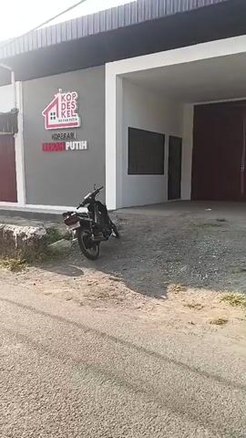 The video shows an exterior view of a building with a sign that reads "KOP DESA KELURAHAN". A motorcycle is parked in front of the building. The camera pans to the right, showing a row of identical stalls or shopfronts with red doors. The road in front of the building is paved with asphalt and appears to be slightly damaged in places. There are some plants growing along the side of the road. Further down the road, there are other buildings, including a multi-story structure. There are no people clearly visible interacting with the motorcycle or the building in the immediate foreground, though figures are seen further down the road. The sky is bright, suggesting daytime. The overall impression is of a commercial or community area in a rural or semi-urban setting.