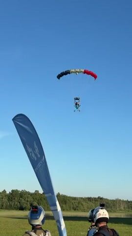 The video shows a tandem skydive landing. Two people are attached to each other, wearing skydiving gear, and are descending under a parachute. The parachute is colorful, with sections of black, red, and white. The skydivers are in the process of landing in a grassy field. In the foreground, a blue flag with a logo is planted in the ground, and three people wearing helmets are looking up at the skydivers. In the background, there are more people gathered, and a small airplane is visible. The sky is clear and blue, indicating good weather conditions. The overall scene depicts the conclusion of a skydive jump.