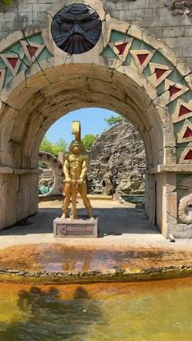 The video shows a golden statue of a warrior holding a sword in front of an arched stone structure. The archway is decorated with triangular patterns in red and green. Above the statue, a carved mask with a fierce expression is visible. Behind the statue, there are rocks and foliage, and a body of water in the foreground. A boat carrying people is visible in the lower right portion of the frame, moving towards the statue. The scene appears to be an outdoor attraction, possibly a theme park or historical reenactment site.