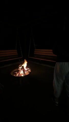 A young person is standing near a fire pit on a dark outdoor deck. The fire pit contains a fire made of wood. The person is wearing a dark long-sleeved shirt and light striped pants. They are standing between two swing benches and gesturing with their hands. The camera angle is slightly above the ground, capturing most of the person’s body, the fire pit, and the swing benches. The environment is very dark, except for the light from the fire.