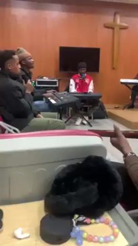 Inside a room with wood-paneled walls and a wooden cross, four people are engaged in a music session. Three are seated at keyboards and one is seated nearby singing into a microphone. A television is in the background, and a fifth person sits on the left side of the screen, filming the musicians. The person filming has a table next to them with headphones, a small round speaker, and colorful beads on it.