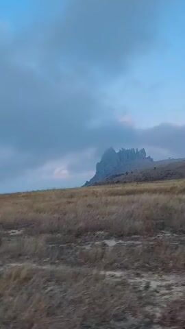 The video shows a wide shot of a dry, grassy field with a distinctive rocky mountain formation in the distance. The sky is overcast with clouds. The camera appears to be moving, creating a slight blur in the foreground grass, suggesting it might be from a vehicle. The mountain is jagged and appears to be a prominent geological feature. The lighting suggests it is either early morning or late evening, with a soft, diffused light. There are no people or other objects clearly visible in the frame.