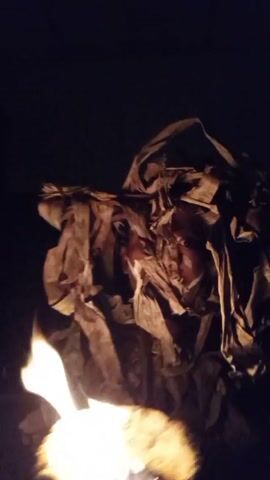 A dark, seemingly nighttime video depicts a person partially concealed behind a mask of brown, shredded paper or cloth, standing near a small fire. The person's eyes are visible through openings in the mask. The flickering light from the fire illuminates the person and the surrounding area. The camera is slightly shaky, adding to the unsettling atmosphere.