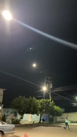 A nighttime video shows a bright moon in a hazy sky. A streetlamp with a bright yellow glow is visible in the lower right corner of the frame, and a utility pole with wires runs vertically on the right side. There are faint diagonal light streaks across the bottom left of the image, possibly from other lights or atmospheric conditions.